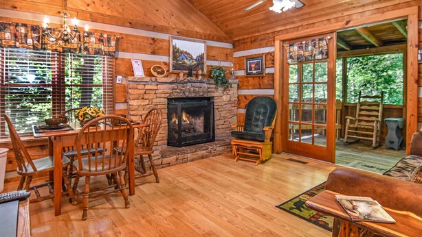 L’intérieur chaleureux d’un chalet avec une cheminée en pierre, des meubles en bois et une grande fenêtre donnant sur un paysage verdoyant.