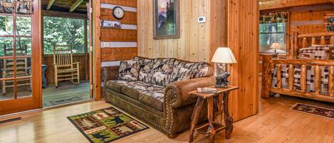 A cozy cabin interior with a wooden floor, a patterned sofa, a small table with a lamp, and a wooden bench with animal prints.