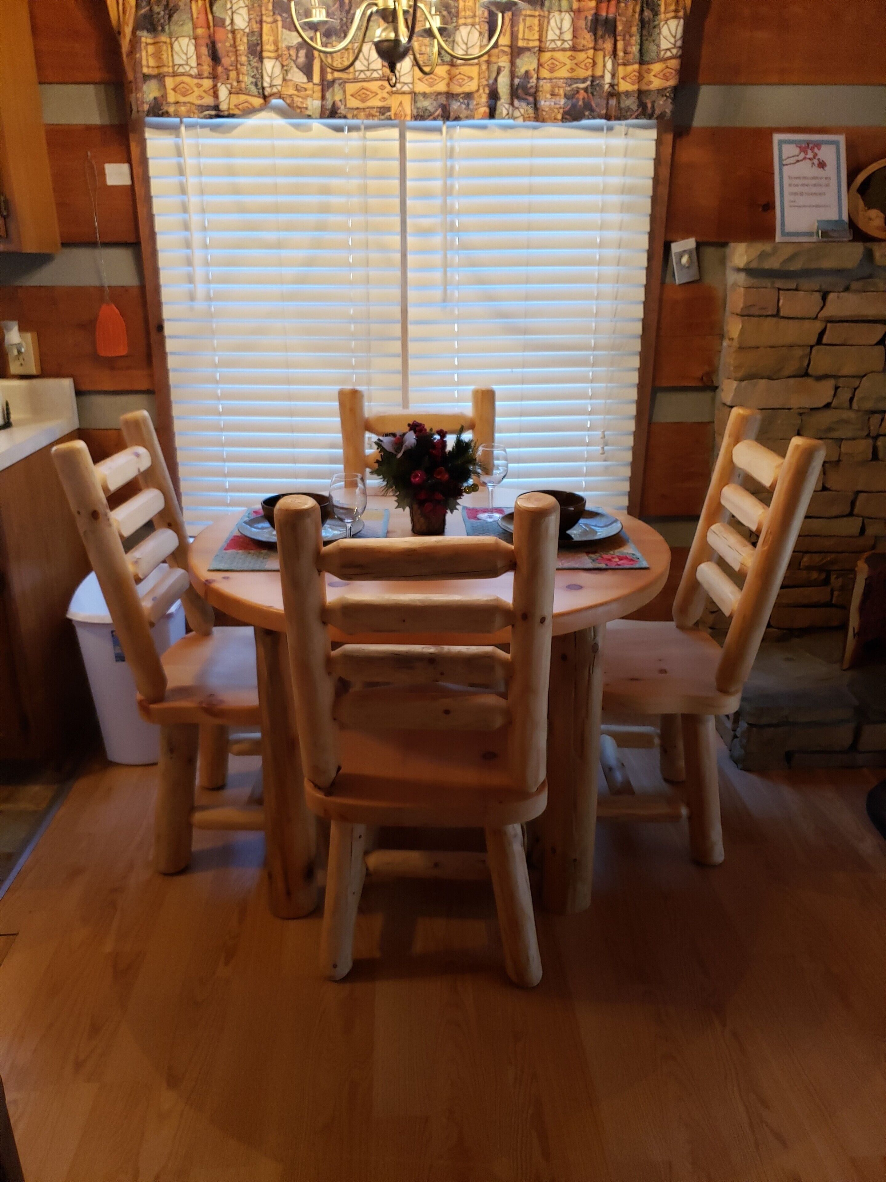 A wooden dining table with four chairs, a vase of flowers, and a stone fireplace in the background.