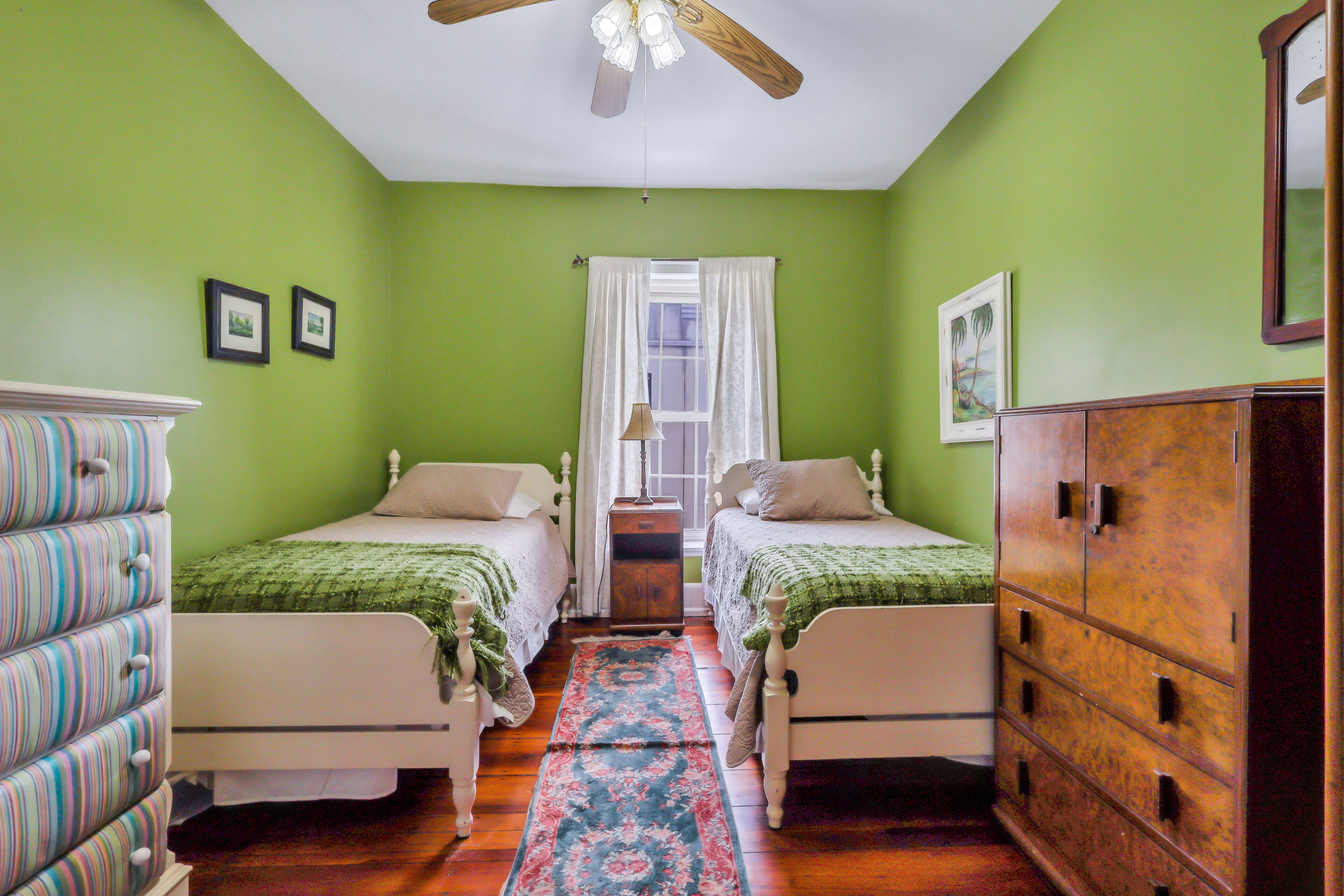 A room with two beds, a wooden dresser, a window with curtains, and a ceiling fan.
