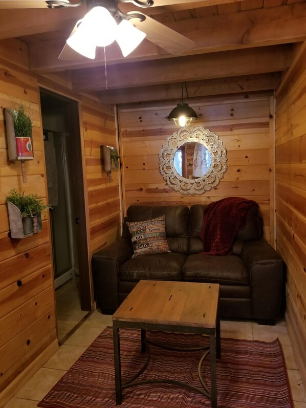 A cozy cabin interior with a wooden interior, a leather sofa, a small table, a mirror, and a ceiling fan. - Charming Lakefront Studio Cabin w/ Private Boat Launch & Slip Ideal for 2 Guests (Westwood)
