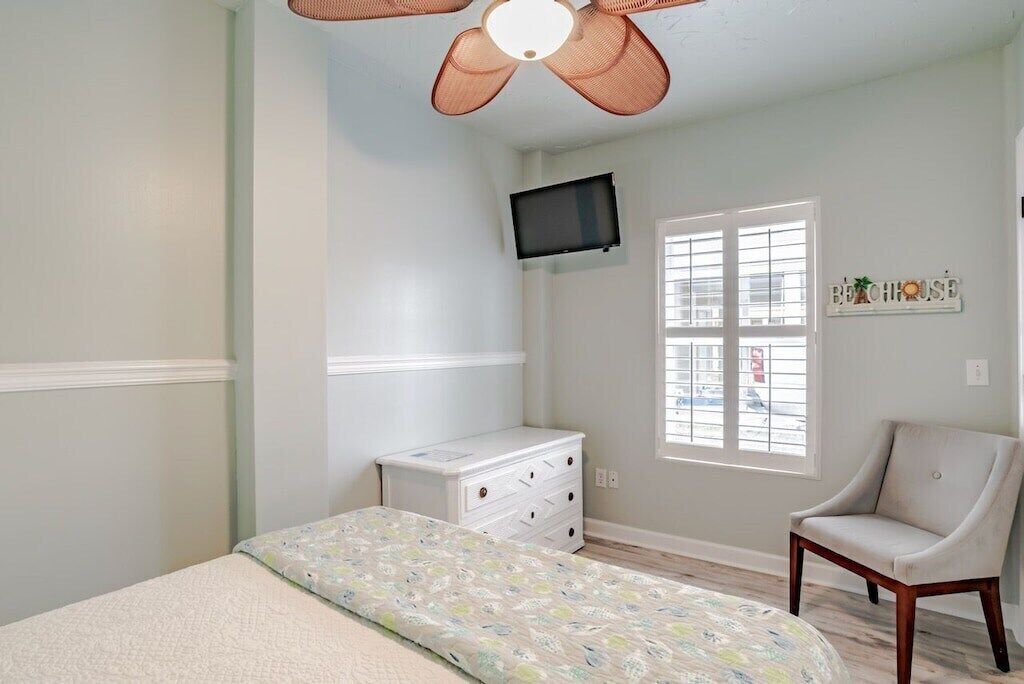 Une chambre à coucher comprenant un lit, une commode, une chaise, un ventilateur de plafond, un téléviseur et une fenêtre.