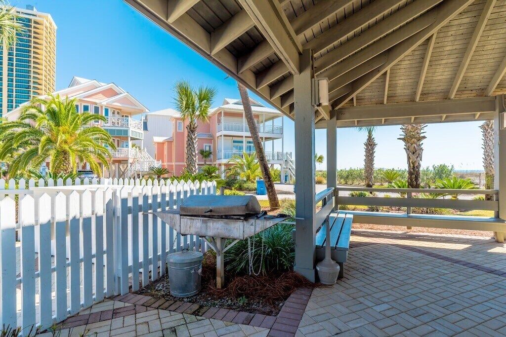 A covered outdoor area with a grill, palm trees, and a white picket fence.