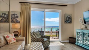 Un salon avec un canapé, un banc, une lampe et des œuvres d’art accrochées aux murs. Il y a un balcon avec vue sur l’océan.