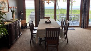 Un comedor con mesa de madera, sillas y una araña. Se aprecia vista al exterior a través de amplios ventanales.