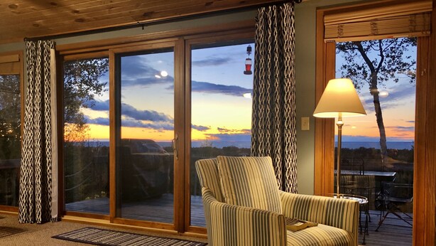 Habitación acogedora con un sillón a rayas, una mesa y una lámpara. Amplias puertas de vidrio dan a una terraza con vistas a la puesta de sol y árboles.