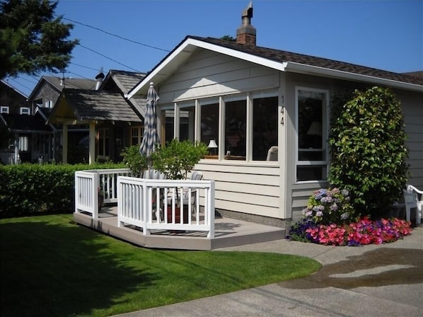 Ein eingeschossiges Haus mit einer überdachten Veranda, einem kleinen Garten mit bunten Blumen und einem weißen Zaun.