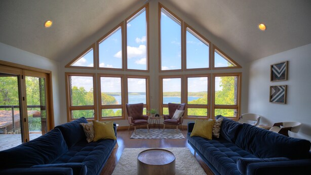 Un vaste salon doté d’un haut plafond et de grandes fenêtres offrant une vue sur un lac et des arbres. On y trouve deux canapés bleu foncé, une table basse ronde et deux fauteuils.