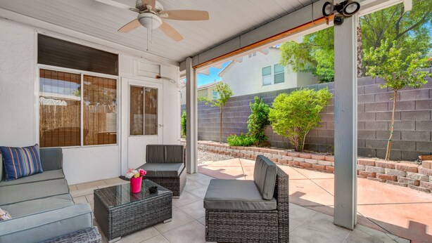A covered patio area with wicker furniture, a ceiling fan, and a view of a brick wall and greenery. - Chic Single Story 3br House Close To Strip & Airport (Las Vegas)