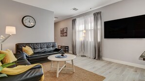 A modern living room with a black leather sofa, a round white coffee table, a wall clock, a flat-screen TV, and a large window with curtains. - Chic Single Story 3br House Close To Strip & Airport (Las Vegas)