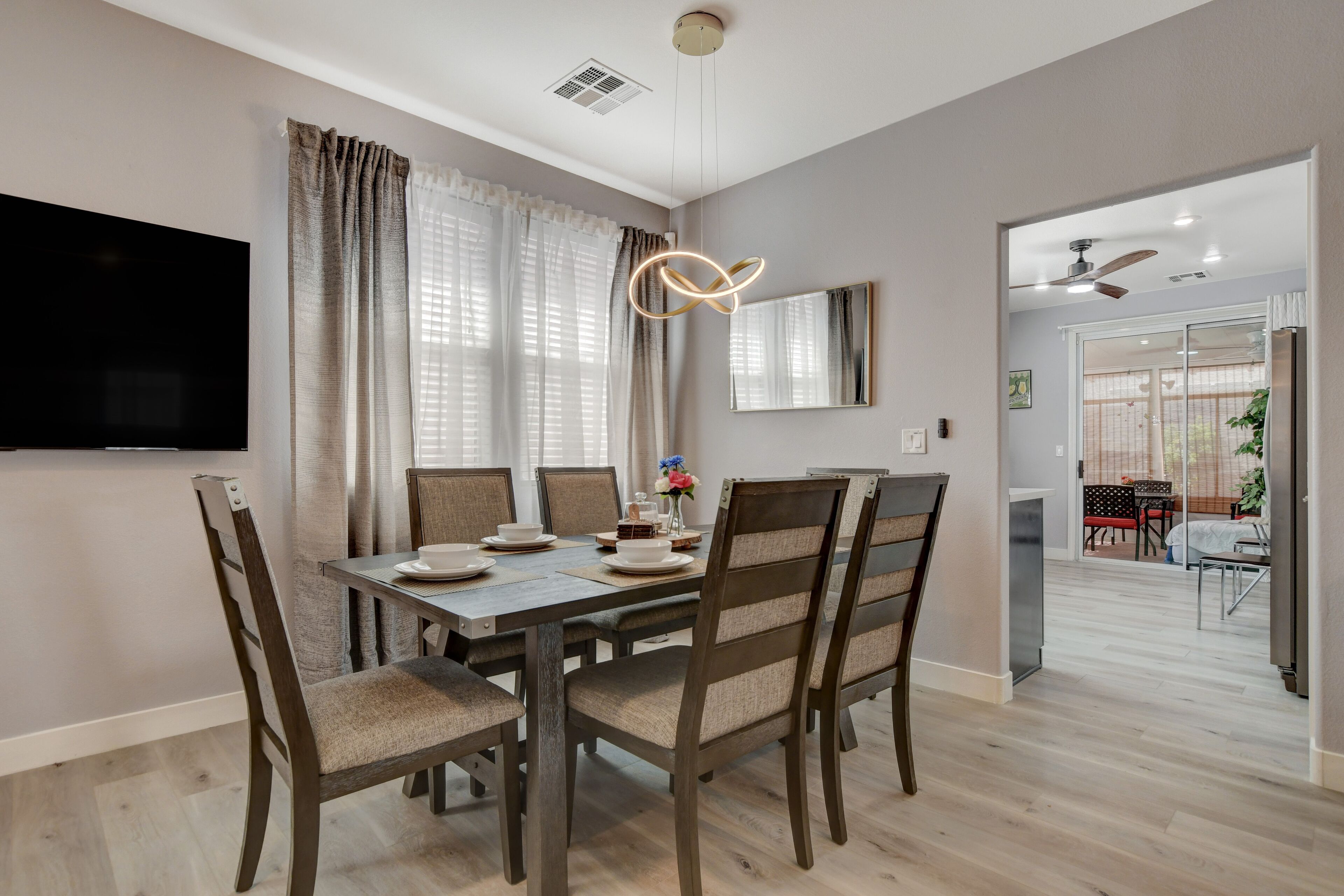 A modern dining area with a wooden table and chairs, a flat-screen TV mounted on the wall, and a ceiling fan.