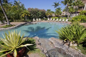 Un área de piscina con sillones de descanso, palmeras y una maceta roja con una planta grande.