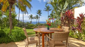 A round wooden table with wicker chairs, surrounded by tropical plants and a view of the ocean.