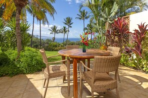 Una mesa de madera redonda con sillas de mimbre, rodeada de plantas tropicales y vista al océano.
