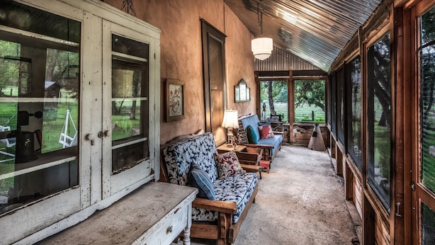 A rustic porch with wooden furniture, a white door, and large windows overlooking a garden.