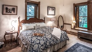 A bedroom with a large bed, wooden headboard, two bedside tables, a nightstand, a mirror, and a window with a view of trees.