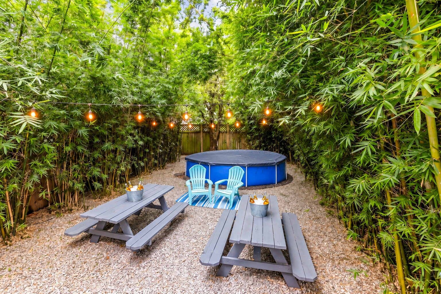 An outdoor seating area with two picnic tables, a blue Adirondack chair, and a hot tub surrounded by bamboo.