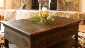 A stone fireplace with a framed painting above it, a glass vase with greenery on a wooden table in front, and a patterned rug on the floor. - Newer Built ski to ski lift , Extra Master Bedroom ,Sleeps 10 (Homewood)