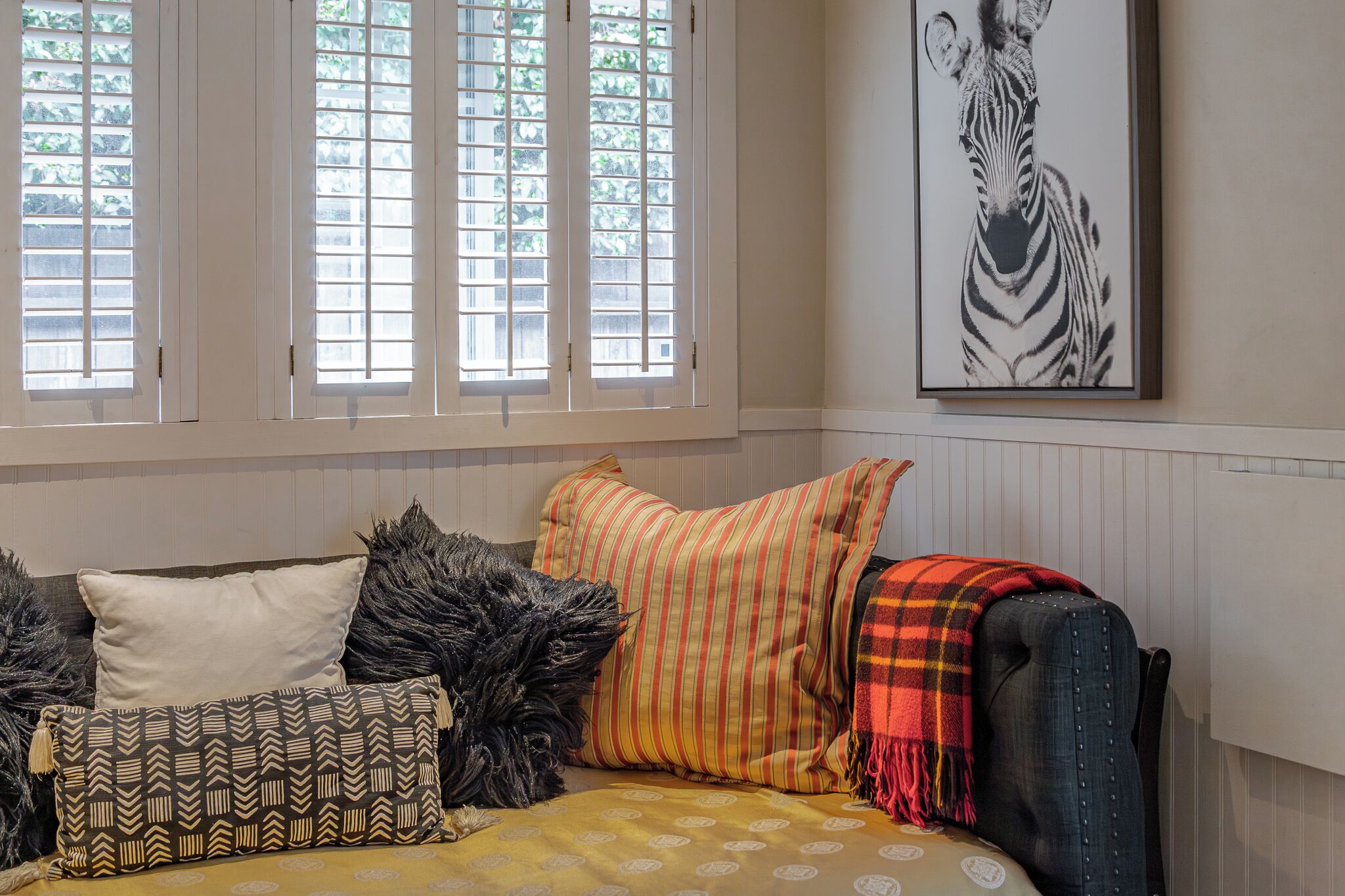 A cozy living room with a sofa adorned with patterned cushions, a zebra artwork on the wall, and a window with blinds.