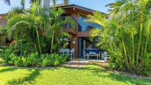 Una casa moderna de dos pisos con balcón, rodeada de abundante vegetación y palmeras.