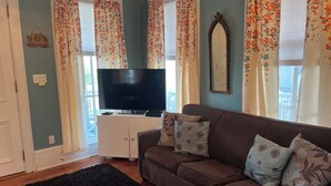 Una sala de estar con un sofá marrón, un televisor de pantalla plana sobre un mueble blanco, cortinas con estampado floral y un espejo grande en la pared.