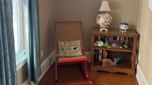 Un pasillo con suelo de madera, una silla roja con un cojín que dice 'BEACH', una pequeña mesita de madera con una lámpara y objetos decorativos, y una ventana con cortinas.