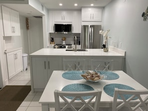 A modern kitchen with white cabinetry, a stainless steel refrigerator, a microwave, and a stove. There is a dining table with blue placemats and a bowl of snacks.