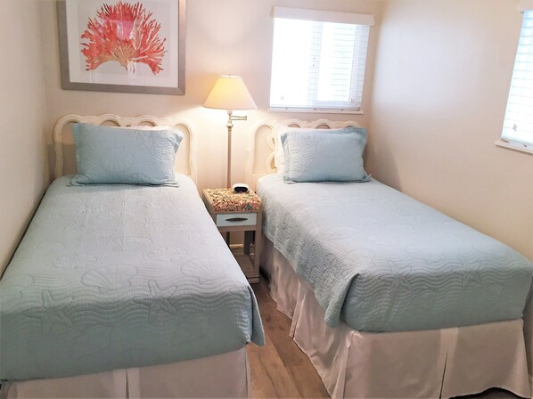 A bedroom with two single beds, a nightstand with a lamp, a framed picture on the wall, and a window with blinds.