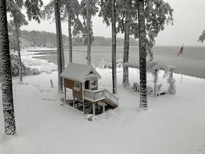 Lumipeitteinen, puinen mökki, jossa on kuisti, ja sen ympärillä on puita ja vesistö.