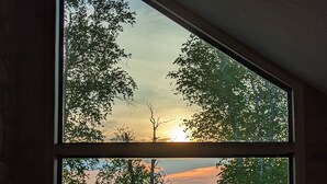 Una habitación con un amplio ventanal con vista a un lago al atardecer, con una terraza de madera con sillas y una mesa.