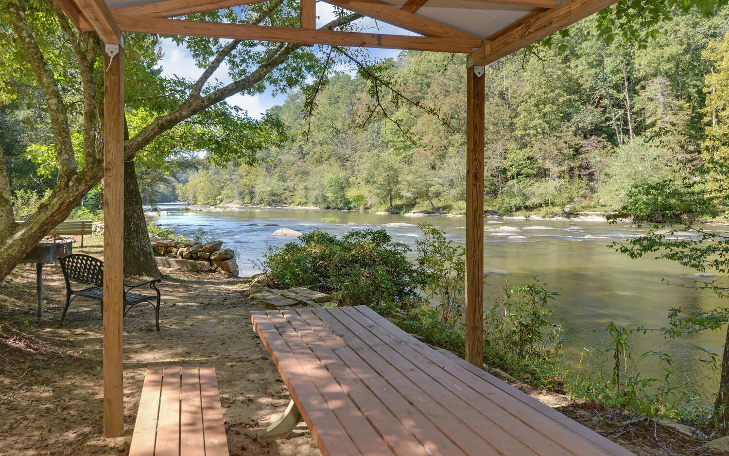 A wooden deck overlooking a river, with a bench and a table under a pergola.