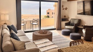 A spacious living room with a large window overlooking a beach, a ceiling fan, and a dining area with wicker furniture. - ALL NEW remodeled condo! Ocean Front on Beach 2 Bd 2 Ba Upper Level (Cabo San Lucas)