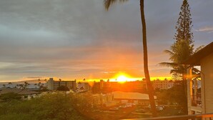 Vista do pôr do sol a partir de uma varanda, com vista para um estacionamento e edifícios, com palmeiras e uma área gramada ao primeiro plano.