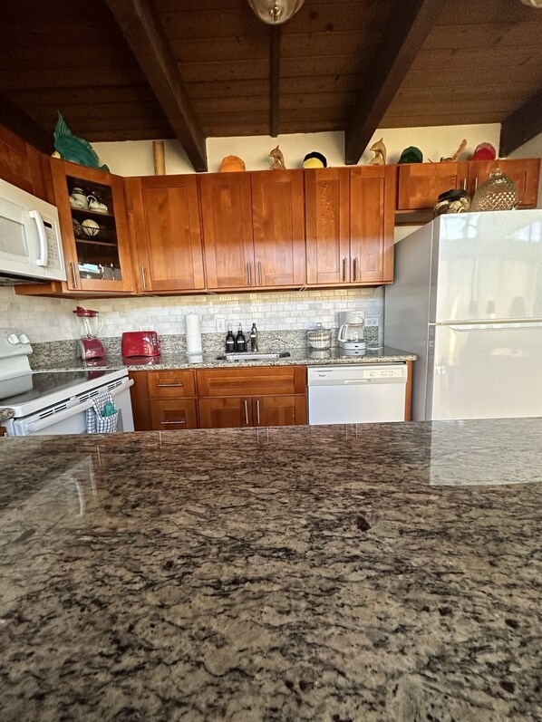 A kitchen with wooden cabinets, a granite countertop, a white refrigerator, and various kitchen appliances. - PARADISE IS CALLING-Oceanview Family Size-Resort Zoned-King Beds-No Cleaning Fee (Kihei)