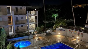 Uma área de piscina à noite, com espreguiçadeiras e um edifício com várias varandas.