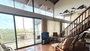 Uma sala de estar ampla com piso de madeira, um ventilador de teto, uma grande janela com vista para palmeiras, um sofá, uma mesa de centro e uma escada.