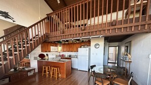 Uma cozinha espaçosa com piso de madeira, uma ilha central com banquetas, uma área de refeições com mesa e cadeiras redondas e uma escada de madeira que leva a um andar superior.