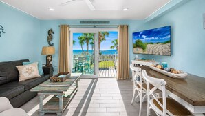 Ein Wohnzimmer mit einer Couch, Couchtisch und Fernseher. Ein Balkon bietet Blick auf Palmen und das Meer.