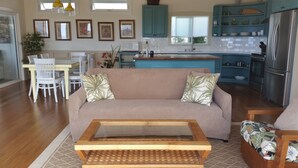 A living room with a beige sofa, a glass coffee table, and a floral armchair. A dining area with a white table and chairs is visible in the background. - Ocean Beach Home In The Heart Of Authentic Old Hawaii (Captain Cook)