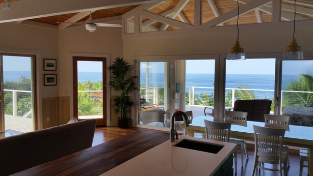 A modern kitchen with a large island, white chairs, and a view of the ocean. - Ocean Beach Home In The Heart Of Authentic Old Hawaii (Captain Cook)