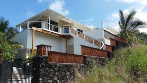 A two-story house with a balcony, a stone wall, and a wooden fence. - Ocean Beach Home In The Heart Of Authentic Old Hawaii (Captain Cook)