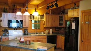 A kitchen with wooden cabinets, a black refrigerator, a microwave, a stove, and a sink. There are bar stools and a window with curtains. - Adventurewood Luxury Log Cabin - Hot Tub, Fireplace, Pool Table (Bloomington)