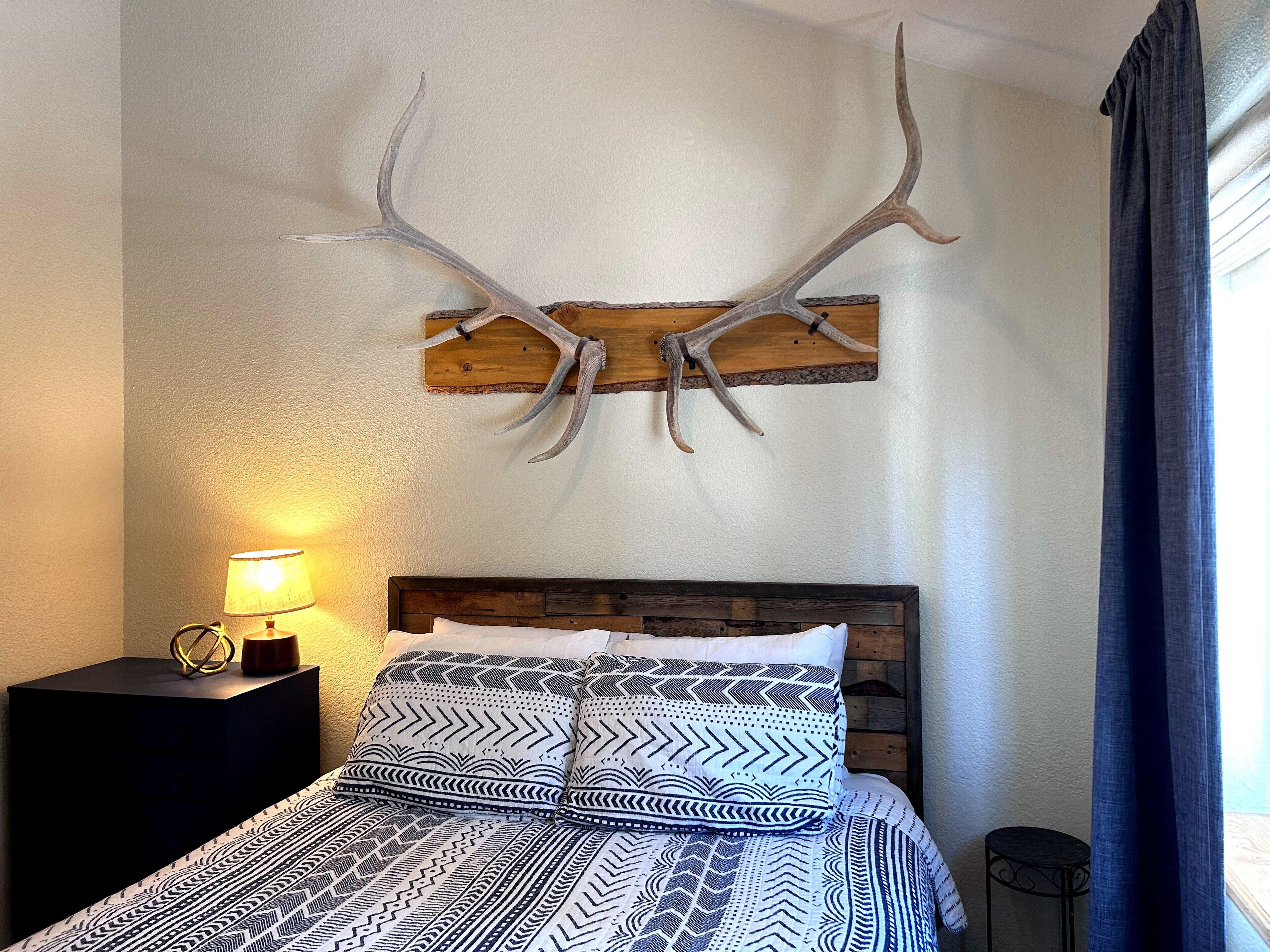 Un dormitorio con cama, mesita de noche, lámpara, decoración de cuernos de ciervo en la pared y ventana con cortinas.