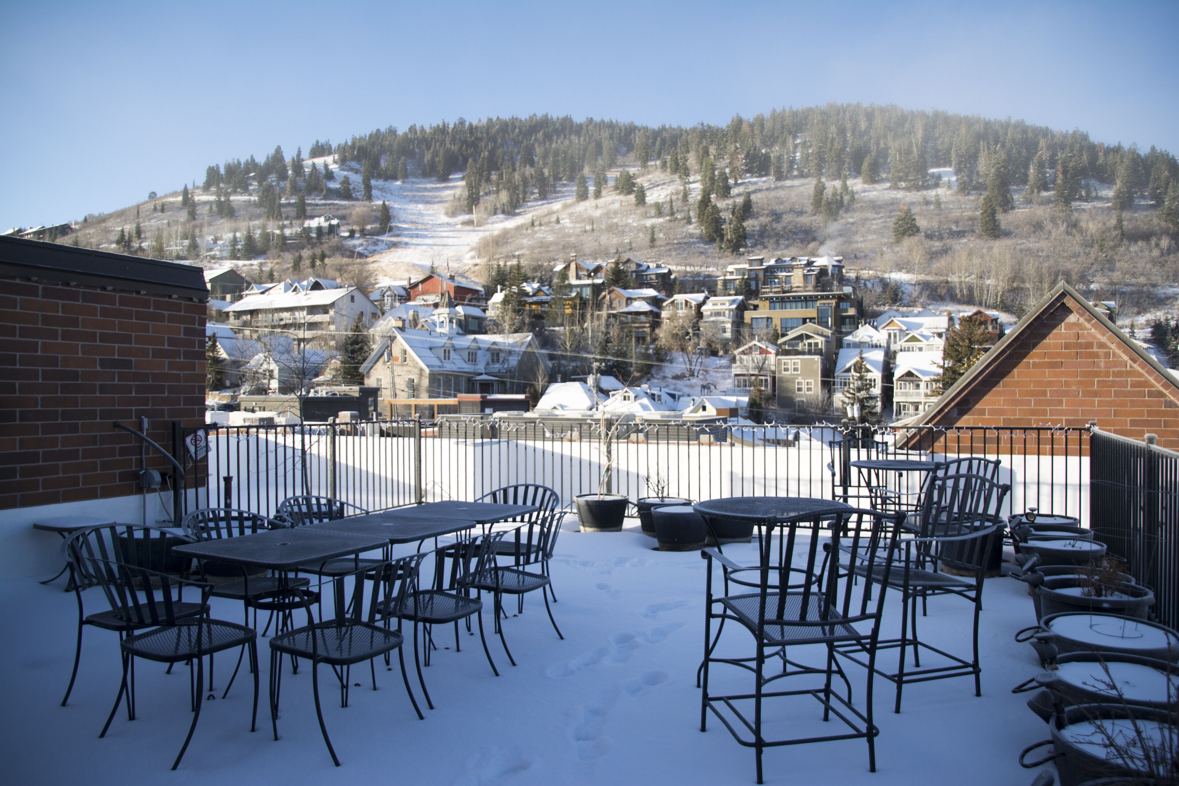 A patio with metal chairs and tables, a brick wall, and a view of a snow-covered village with houses and a hill in the background.
