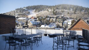 Un patio con sillas y mesas de metal, un muro de ladrillo y una vista de un pueblo cubierto de nieve con casas y una colina al fondo.