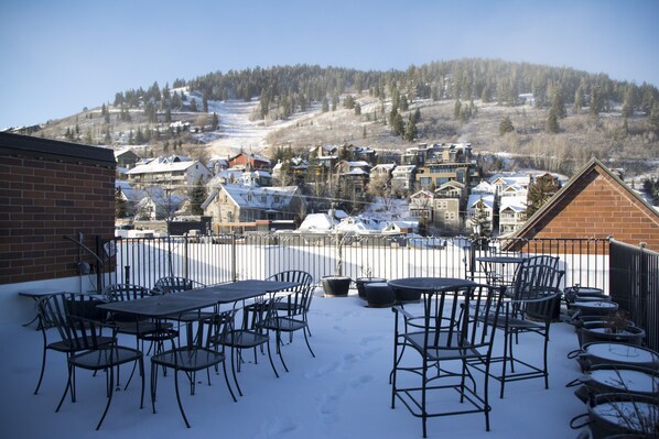 A patio with metal chairs and tables, a brick wall, and a view of a snow-covered village with houses and a hill in the background.