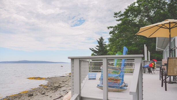 A wooden deck with a railing overlooking a beach, a blue chair, and a yellow umbrella.