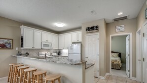 Una cocina con armarios blancos, encimera de granito y refrigerador. Hay taburetes de madera en la encimera.