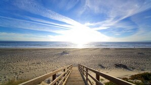 Una pasarela de madera que lleva a una playa, con cielo despejado y el sol poniéndose o saliendo.