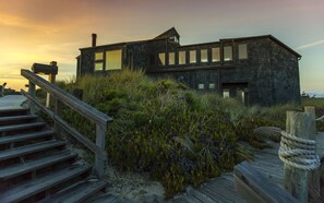 A modern building with large windows and a chimney, surrounded by natural vegetation and wooden stairs leading up to it. - At The Beach! Insanely Great Beach House, Ocean Views, Relaxation & Pickleball (Pajaro Dunes)